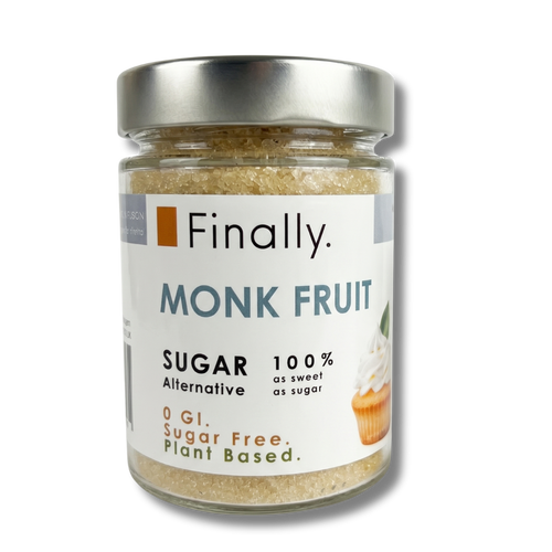 A small glass jar with a metallic silver lid, filled with Finally brand "MONK FRUIT Sugar Alternative", isolated against a transparent checkerboard background. The clear jar reveals a light golden-brown, crystalline sugar-like texture inside. The white label features slate blue text reading "MONK FRUIT", alongside product details including "100% as sweet as sugar", "0 GI. Sugar Free. Plant Based.", and a small graphic of an iced cupcake.