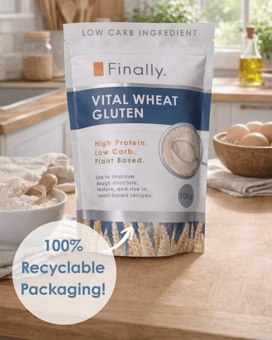 A product shot featuring a 700g stand-up pouch of Finally brand VITAL WHEAT GLUTEN positioned in the centre of a wooden kitchen worktop. The white and blue pouch is labeled as a "LOW CARB INGREDIENT" that is "High Protein. Low Carb. Plant Based." A prominent circular overlay in the bottom left corner includes an arrow pointing to the pouch with the text "100% Recyclable Packaging!". 