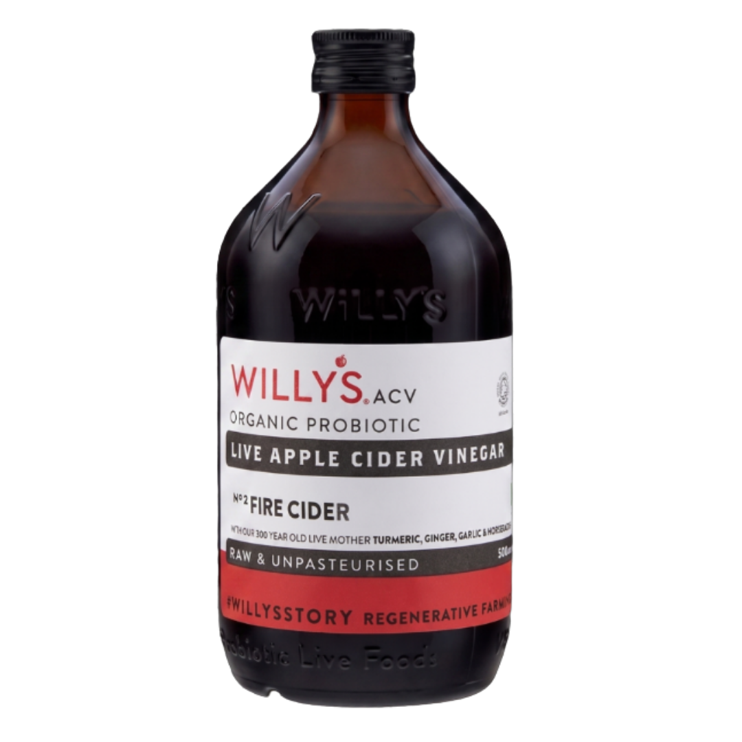 A 500ml amber glass bottle of Willy's ACV "ORGANIC PROBIOTIC LIVE APPLE CIDER VINEGAR", isolated against a transparent checkerboard background. The bottle has a black screw cap and features embossed lettering reading "WILLY'S" on the glass shoulder. The white label indicates it is "Nº2 FIRE CIDER", infused with "TURMERIC, GINGER, GARLIC & HORSERADISH", and states it is "RAW & UNPASTEURISED" with a solid red band across the bottom.