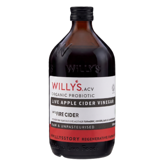 A 500ml amber glass bottle of Willy's ACV "ORGANIC PROBIOTIC LIVE APPLE CIDER VINEGAR", isolated against a transparent checkerboard background. The bottle has a black screw cap and features embossed lettering reading "WILLY'S" on the glass shoulder. The white label indicates it is "Nº2 FIRE CIDER", infused with "TURMERIC, GINGER, GARLIC & HORSERADISH", and states it is "RAW & UNPASTEURISED" with a solid red band across the bottom.